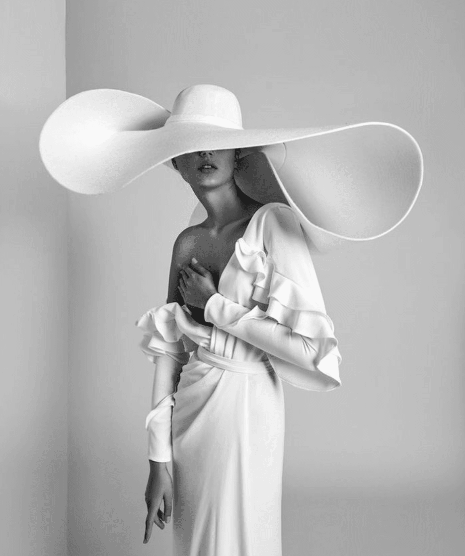 Editorial fashion photograph of a sculptural white oversized hat with flowing dress